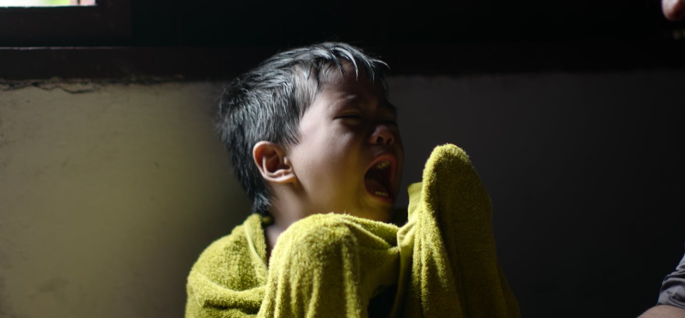 A young boy wrapped in a towel sits crying by a window, showing displeasure.