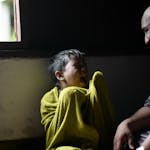 A young boy wrapped in a towel sits crying by a window, showing displeasure.