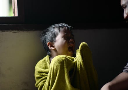 A young boy wrapped in a towel sits crying by a window, showing displeasure.