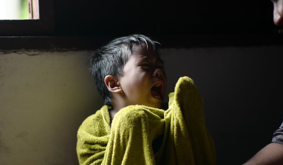 A young boy wrapped in a towel sits crying by a window, showing displeasure.